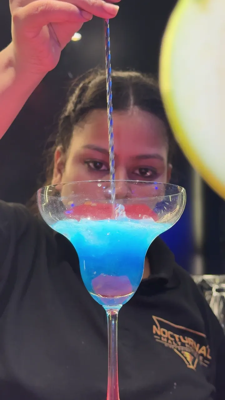 A bartender stirs a layered blue cocktail in a margarita glass