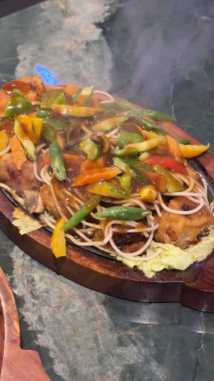 A sizzler with noodles and peppers steaming on a wooden board